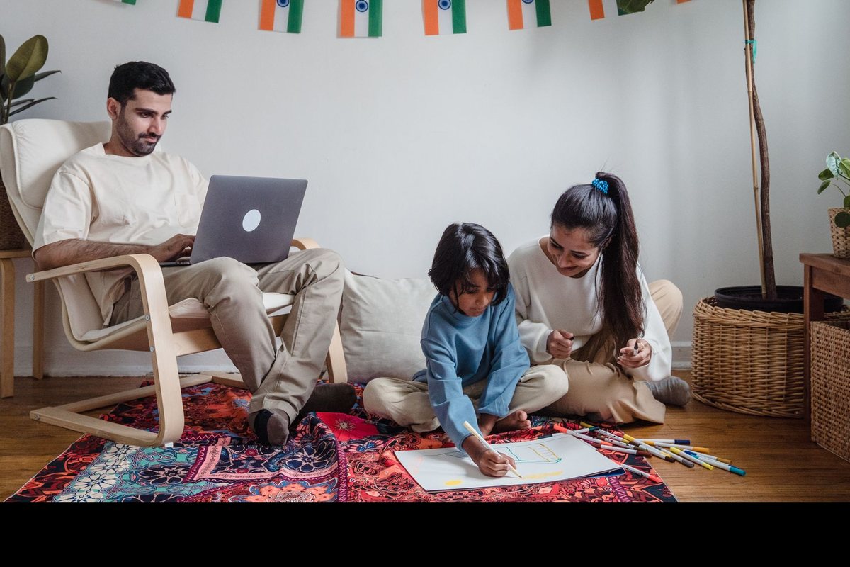 A father on his laptop at home while his two children draw beside him